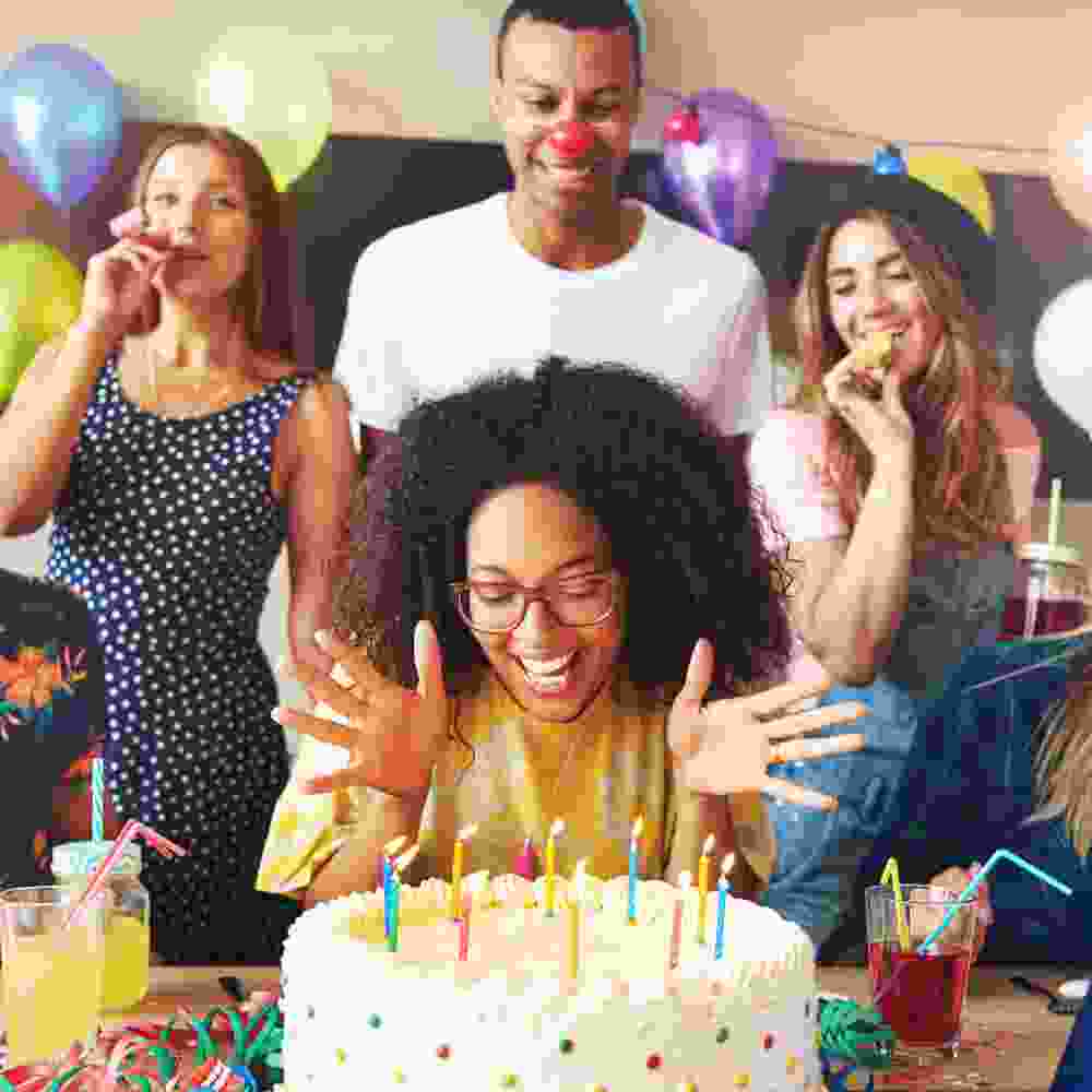 Group celebrating a birthday with a joyful woman in front of a decorated cake, colorful balloons, and drinks, emphasizing a festive dining experience.