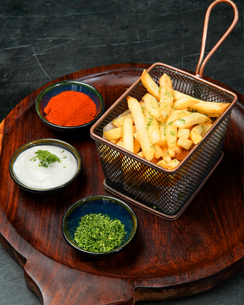 Crispy French fries in a metal basket with bowls of green herbs, creamy dip, and red spice on a wooden platter, highlighting High Brew's fresh and flavorful meal offerings.